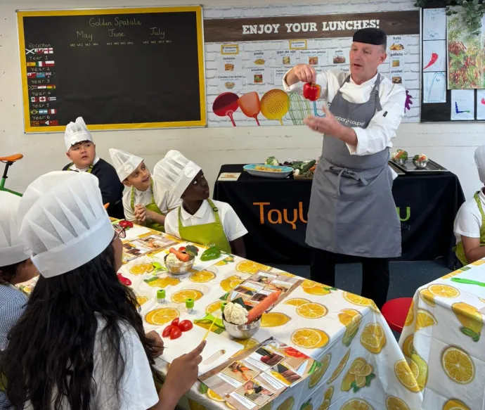 A male chef standing at the front holding a red pepper. At the front our six our pupils sat at a table with a selection of vegetables listening to the chef